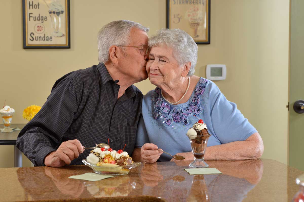 Senior adult man kissing cheek of woman eating sundaes at table
